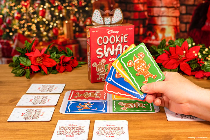 Lifestyle photo of the Disney Cookie Swap card game laid out on the floor in front of a Christmas tree and pointsetta plants