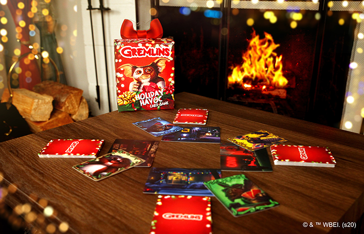 Lifestyle photo of the Gremlins Holiday Havoc card game laid out on a table in front of a roaring fireplace decorated with holiday lights