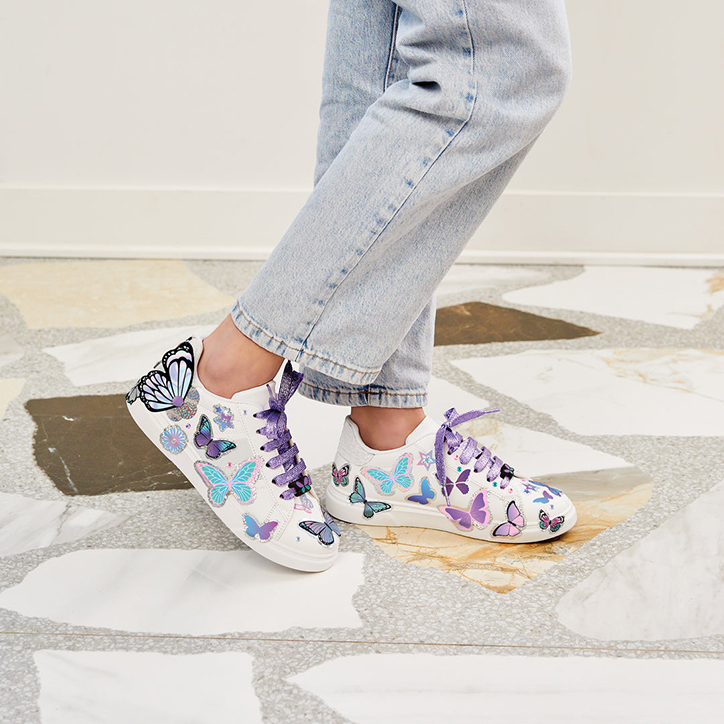 Lifestyle photo of a girl standing outdoors wearing white sneakers with sparkly purple shoelaces, butterfly stickers, and rhinestones designed with the Make It Real Sticker Chic Sneaker Design Kit