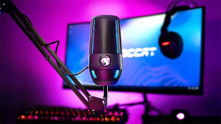 Lifestyle photo of the ROCCAT Torch Studio-Grade USB Microphone sitting in front of a monitor and keyboard. There is a purple glow in the background.