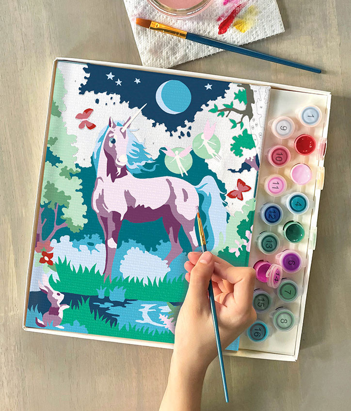 Lifestyle photo of the iHeartArt Paint by Numbers Moonlit Unicorn kit with a near completed painting of a unicorn standing in a moonlit forest with fairies floating all around