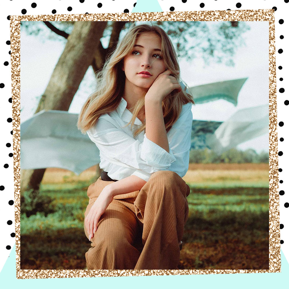 Lacey Caroline sitting outdoors in a white blouse and tan pants. She has her hand on her face and is looking up as if she is daydreaming