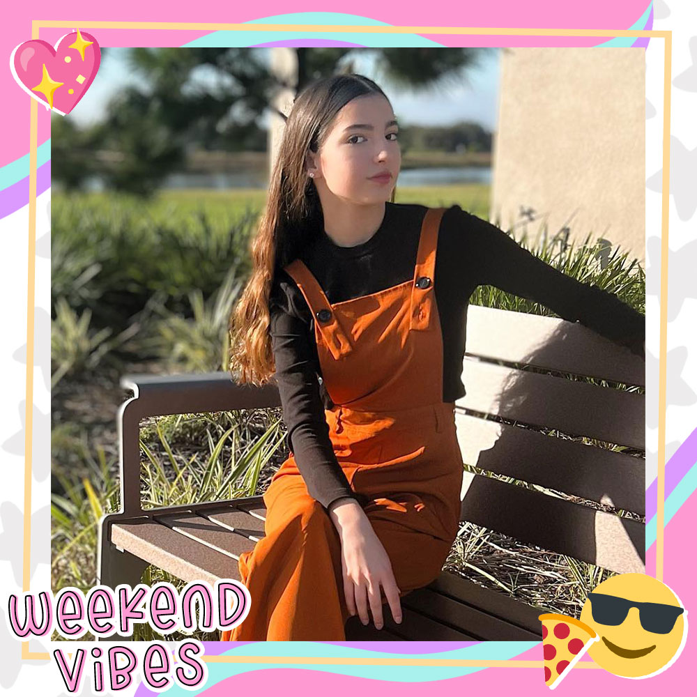 Ariana Nunez sits on a bench outdoors wearing burnt orange overalls and a black long sleeve shirt