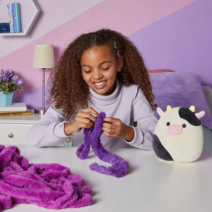 Tween girl sitting in a purple and pink bedroom knitting a scarf using the Big Fat Yarn Squishmallows Accessory Kit