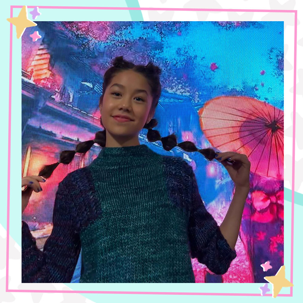 Trinity Jo-Li Bliss wearing bubble braids, standing in front of a backdrop featuring cherry blossoms and a woman wearing a kimono, holding a parasol