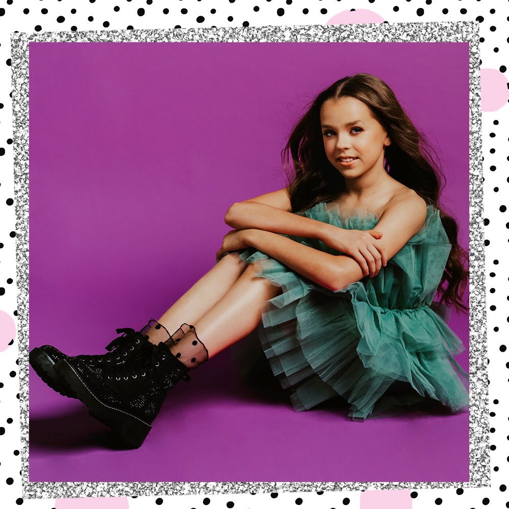 Tatum Brady in a teal ruffly dress, polkadot lace socks, and velvet combat boots sits on the floor in front of a purple backdrop