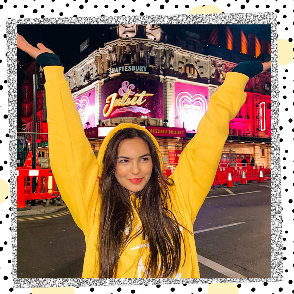 Annissa Murad standing in front of a theater in a yellow hoodie with her hands in the air