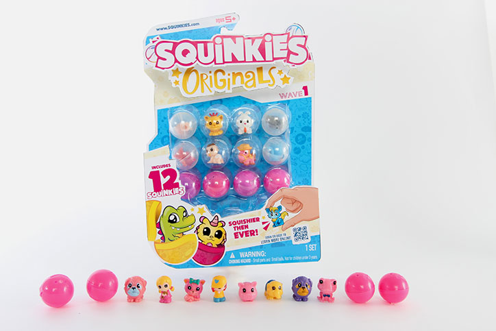 Product image of a Squinkies Originals 12 Pack with a few unboxed Squinkies and pink plastic bubbles lined up in front of the box
