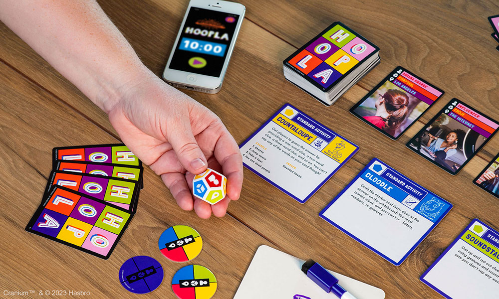 A hand holding a Cranium Hoopla die with cards, game pieces, and a timer laying around a wooden table