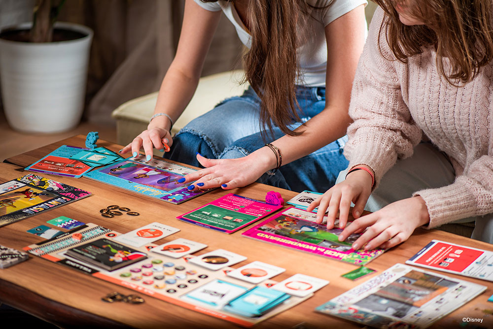 Two teen girls sitting on a couch, assembling their Disney Animated game boards on a table in front of them
