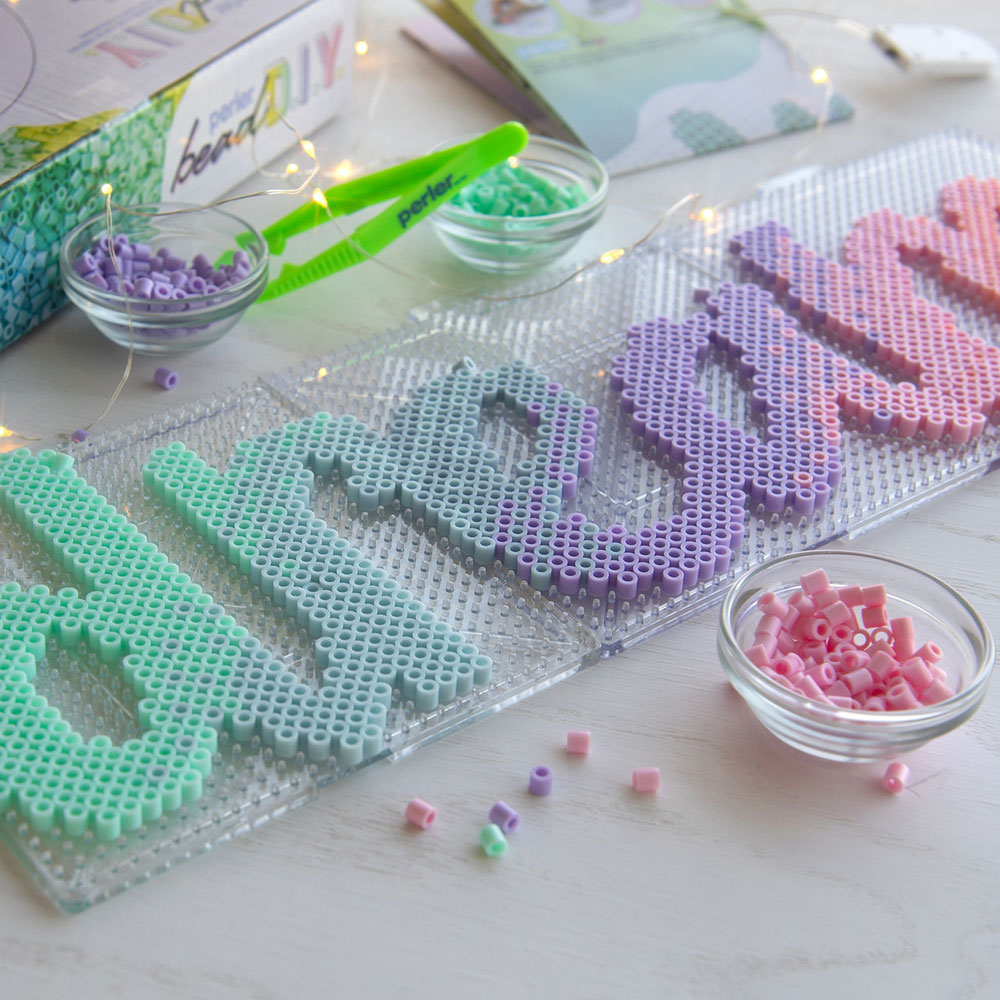 Lifestyle photo of the Bead D.I.Y. Dream Activity Kit from Perler. The word "dream" is written out in pastel Perler. beads across three clear pegboards, surrounded by fairy lights, bowls of beads, and the product box