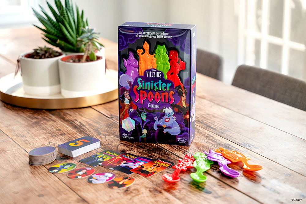 Sinister Spoons game box on a table by a window with light shining in. The game pieces and cards surround the box and three succulent plants can be seen in the background.