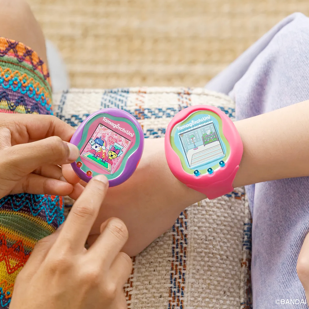 Two kids sit on a couch, each with a Tamagotchi Uni device on their wrists.