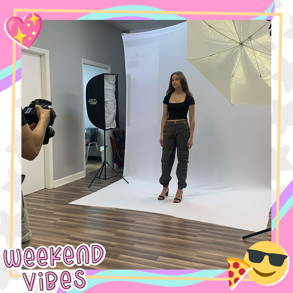 Paige Stein in front of a photoshoot backdrop modeling a black tee and green cargo pants. A photographer is seen off to the side taking her photo.