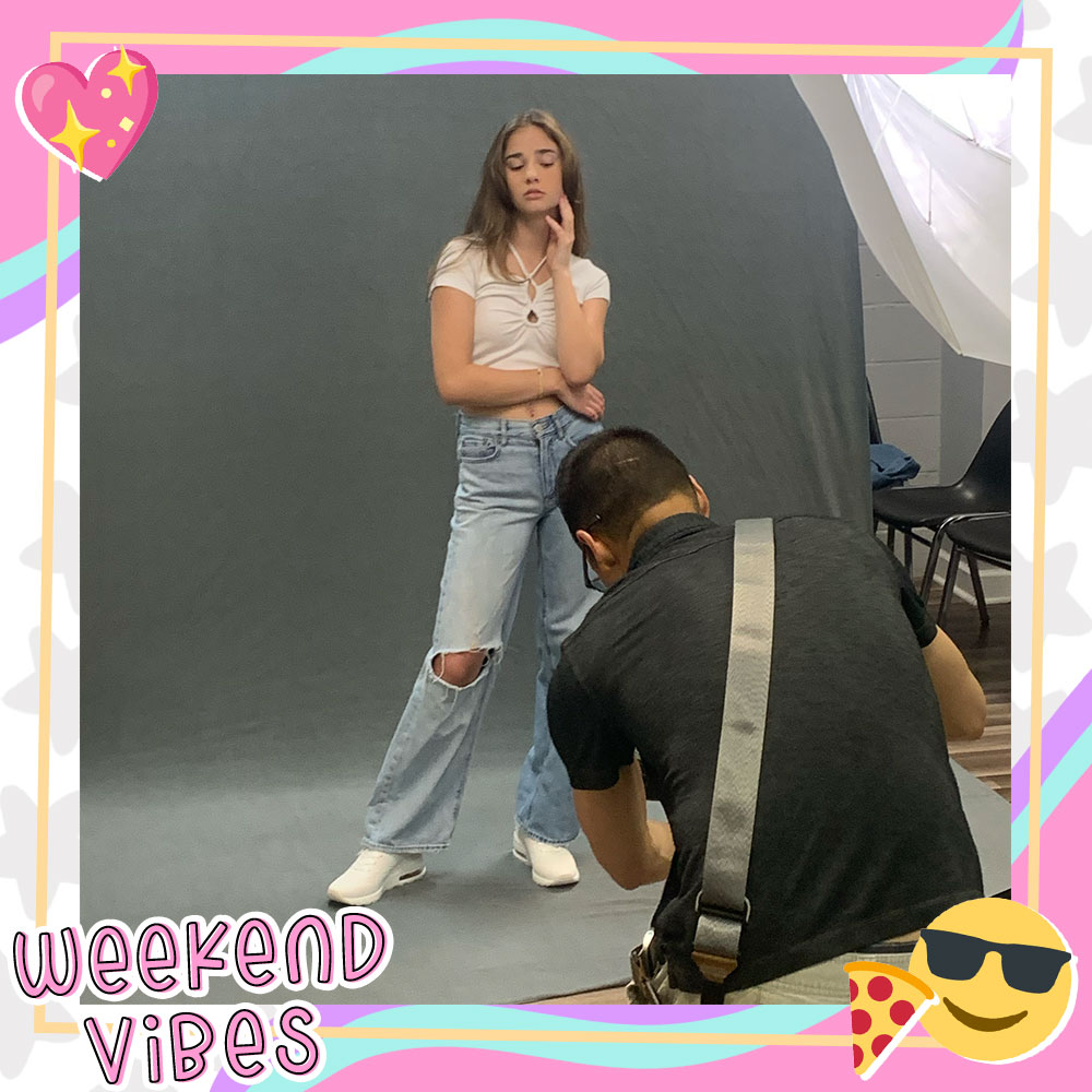Paige Stein in front of a photoshoot backdrop modeling a white tee and baggy ripped jeans. A photographer is seen in front taking her photo.