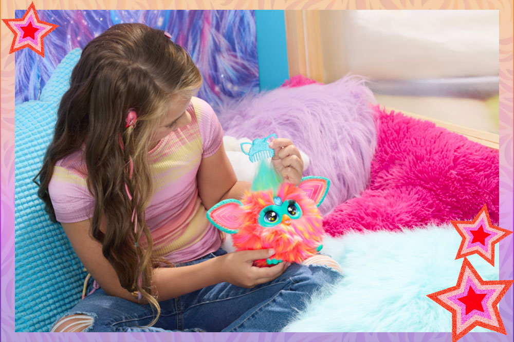 A tween girl sitting in a bed surrounded by colorful pillows. She is holding a coral Furby and combing its hair.