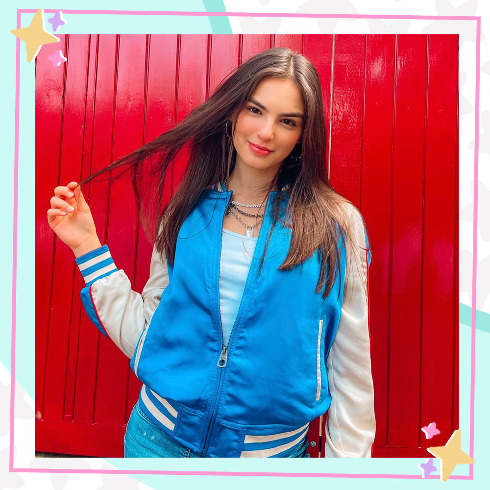 Annissa stands in front of a red fence in a blue and white varsity jacket and layered necklaces. She is holding a strand of her hair and smiling.