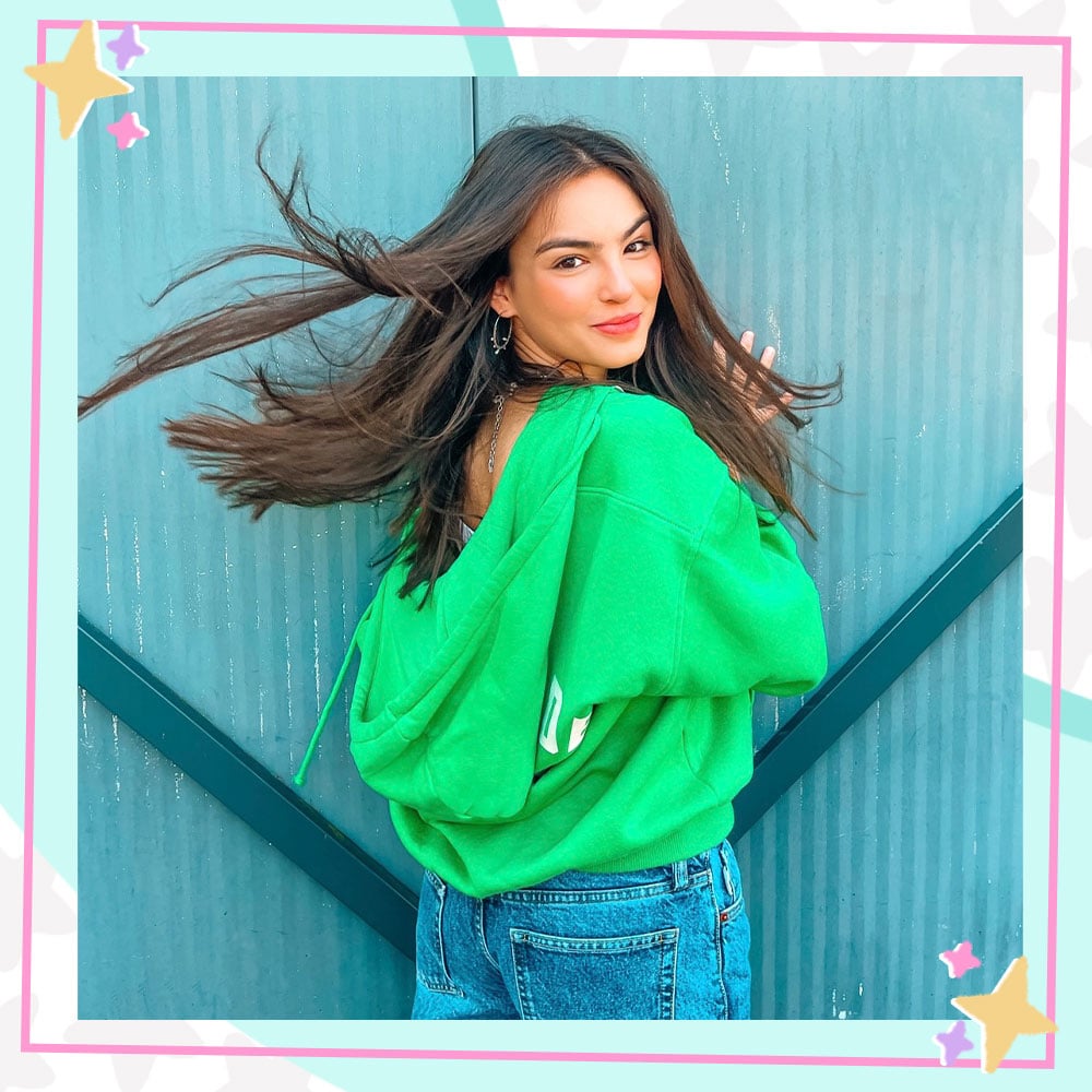 Annissa Murad poses in front of larget metal slats. She is wearing a green hoodie and baggy jeans and is looking over her shoulder at the camera.