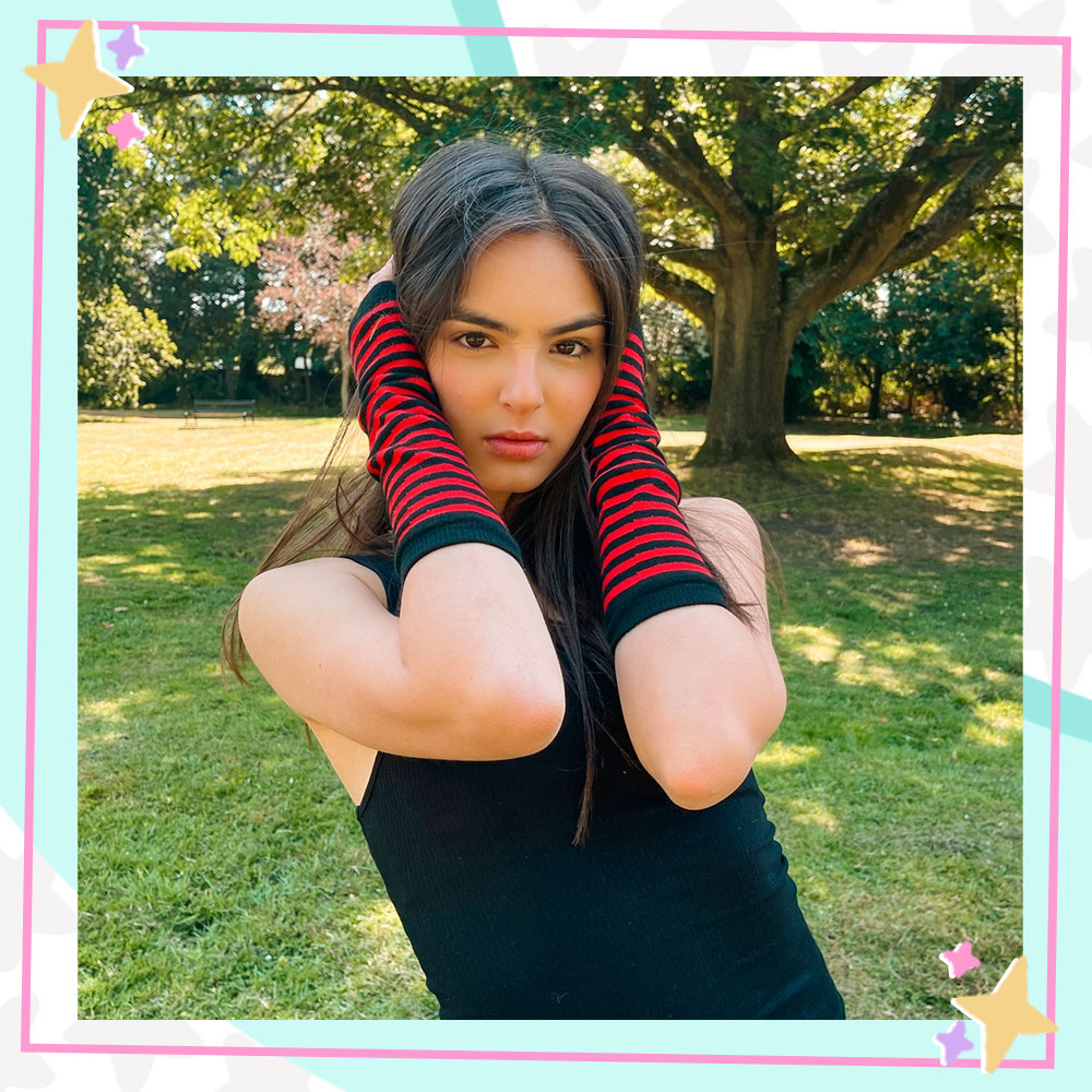 Annissa stands in a grassy field with a tree in the distance. She is wearing a black tanktop and black and red striped armwarmers. Her hands are covering her ears.