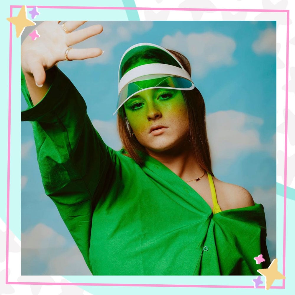 Kenz Couch in front of a cloud backdrop, wearing an off the shoulder green button down top and a green visor, raising her hand.