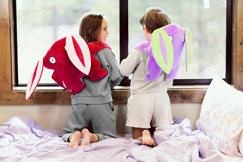 Two kids kneeling on a bed, looking out the window, holding their Bumpas plush