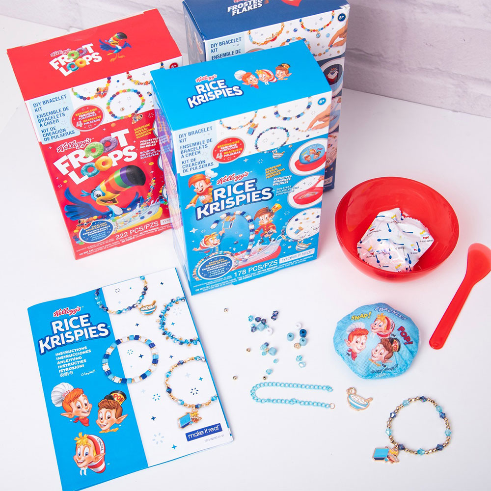 All three Make it Real Kellogg's DIY Bracelet Kits sitting on a kitchen counter. The kit pieces are laid out below them, including the instructions, bracelet cord, beads, cereal bowl, spoon, and dissolvable pouch.