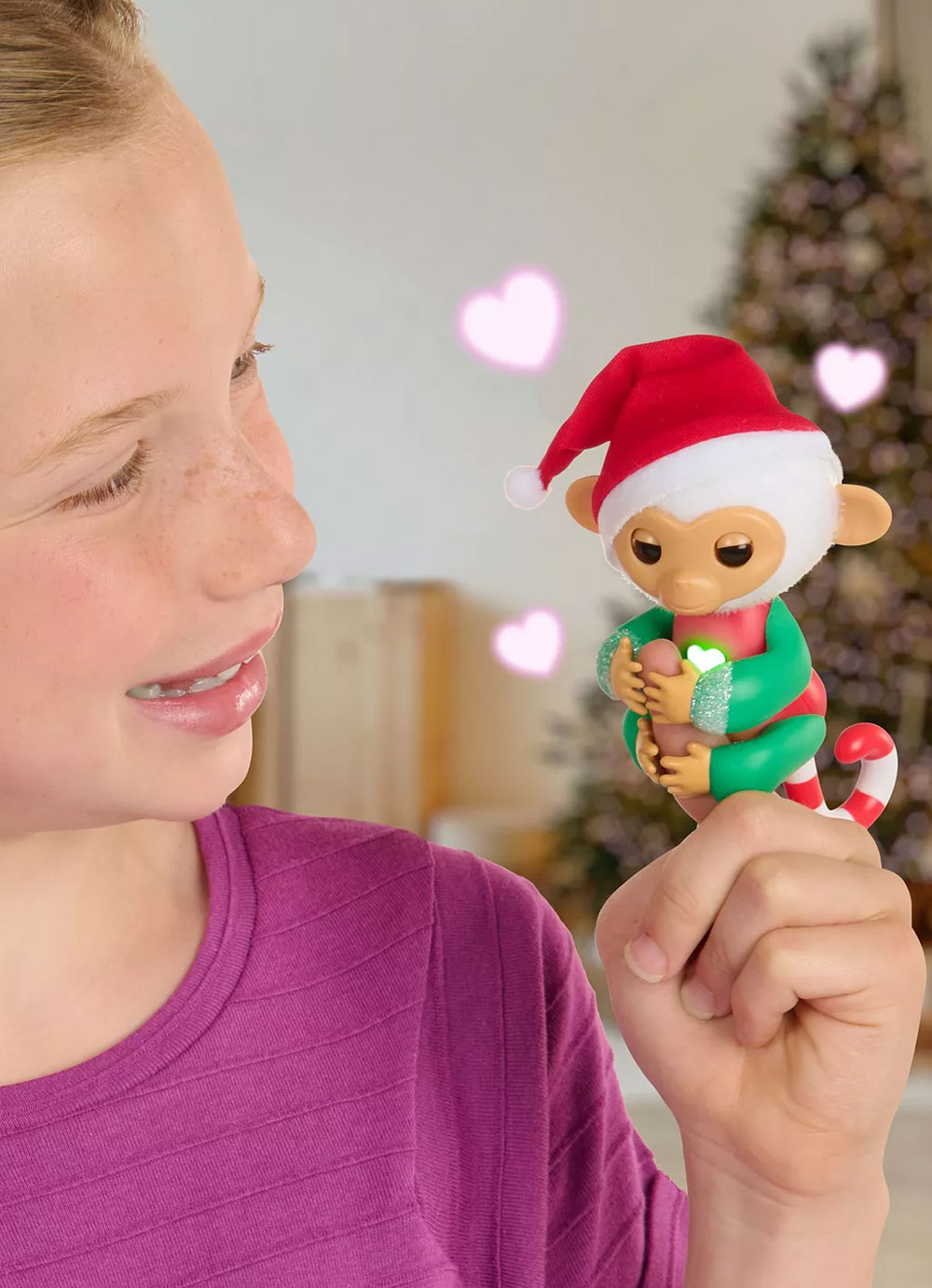A tween girl holding a Snowbelle Fingerlings baby monkey on her finger in front of a Christmas tree