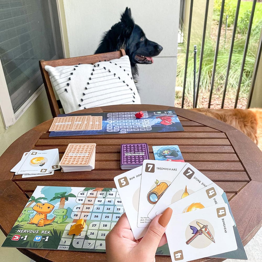 A person sits at an outdoor table with the Happy Little Dinosaur boards and cards spread out on the table. There is a dog in the background.