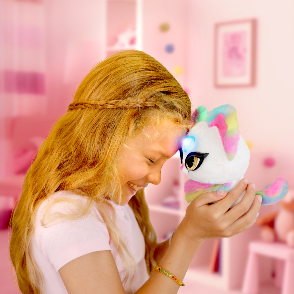 A tween girl standing in a pink bedroom holding her Magic Whispers Kitty up to her head to her the secret message