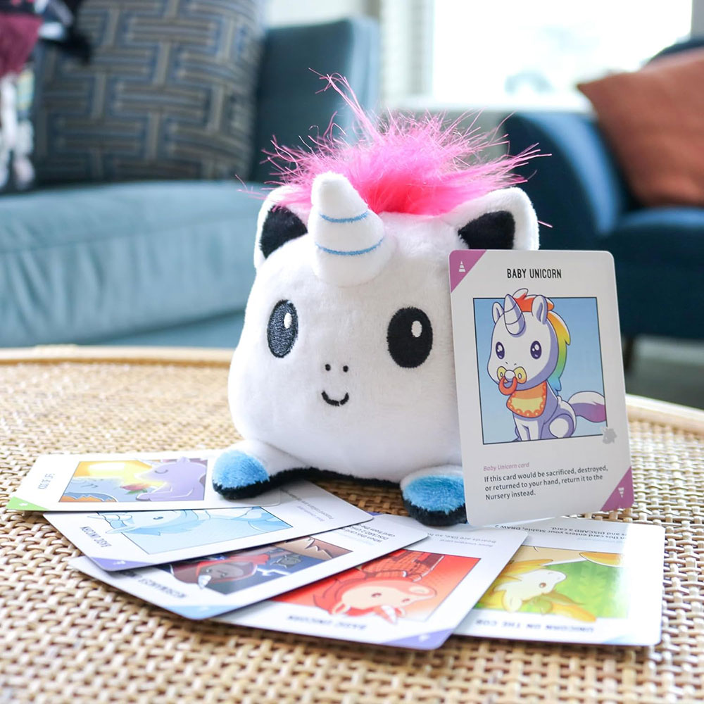 A plush unicorn sits on a table, surrounded by Unstable Unicorns game cards