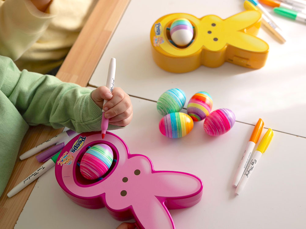 A kid decorating eggs using a pink PEEPS Eggmazing Egg Decorator. There are colored eggs and markers scattered around the table.